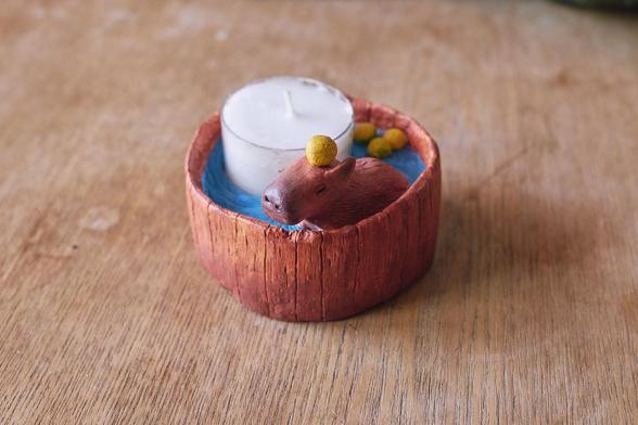 A small candle holder made of clay, representing a capybara bathing in a round wooden baththub with yuzu fruits